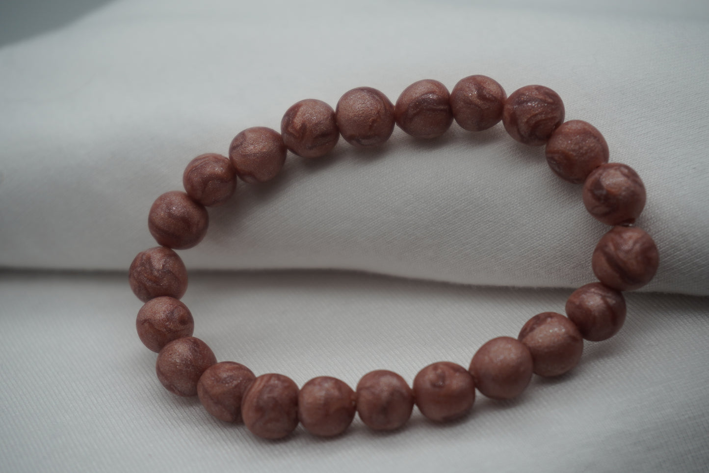 A bracelet made of rose gold-colored round beads against a white backdrop.  The beads sparkle and have a slight swirled pattern.
