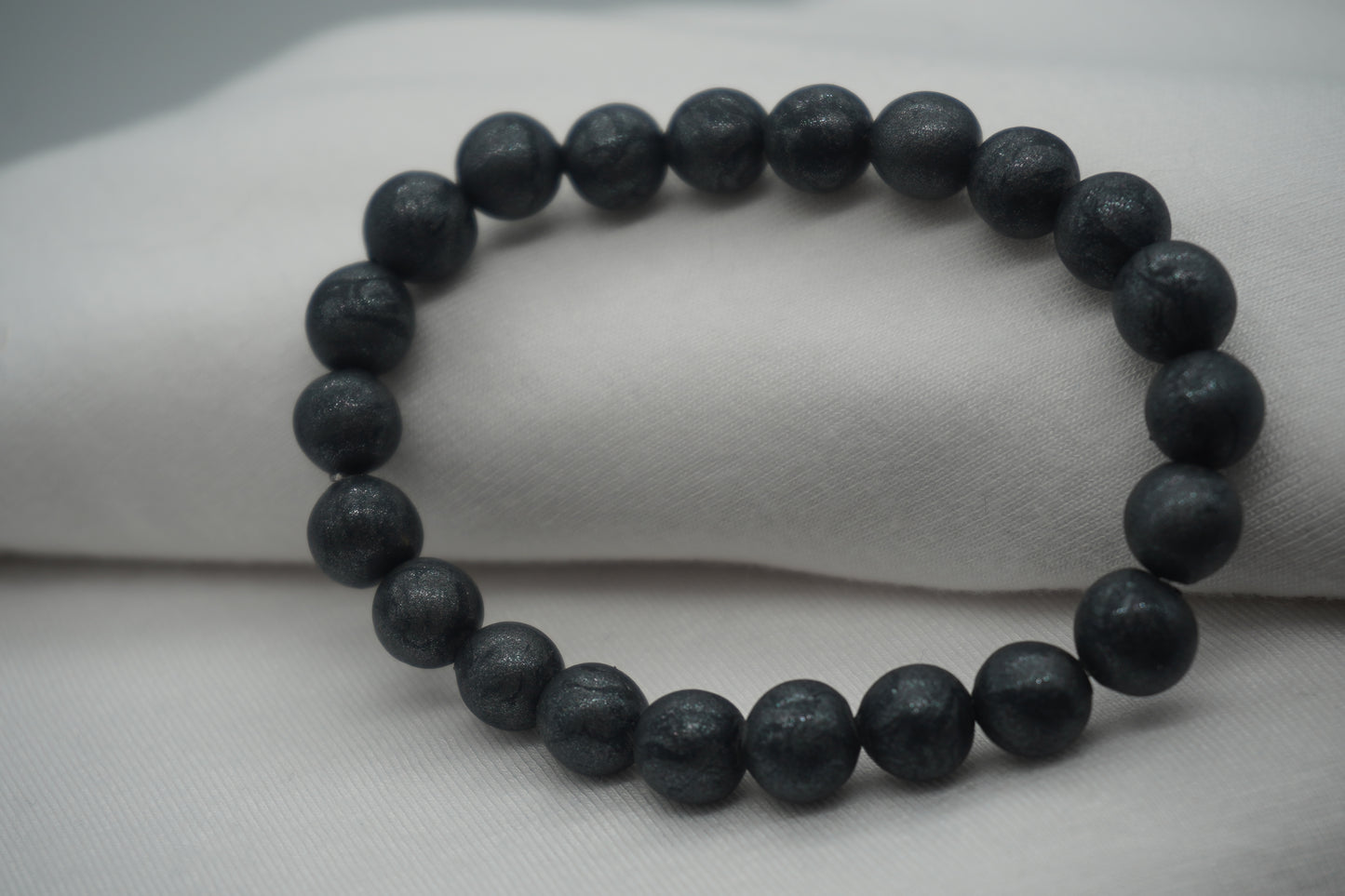 A bracelet made of dark grey-colored round beads against a white backdrop.  The beads sparkle and have a slight swirled pattern.