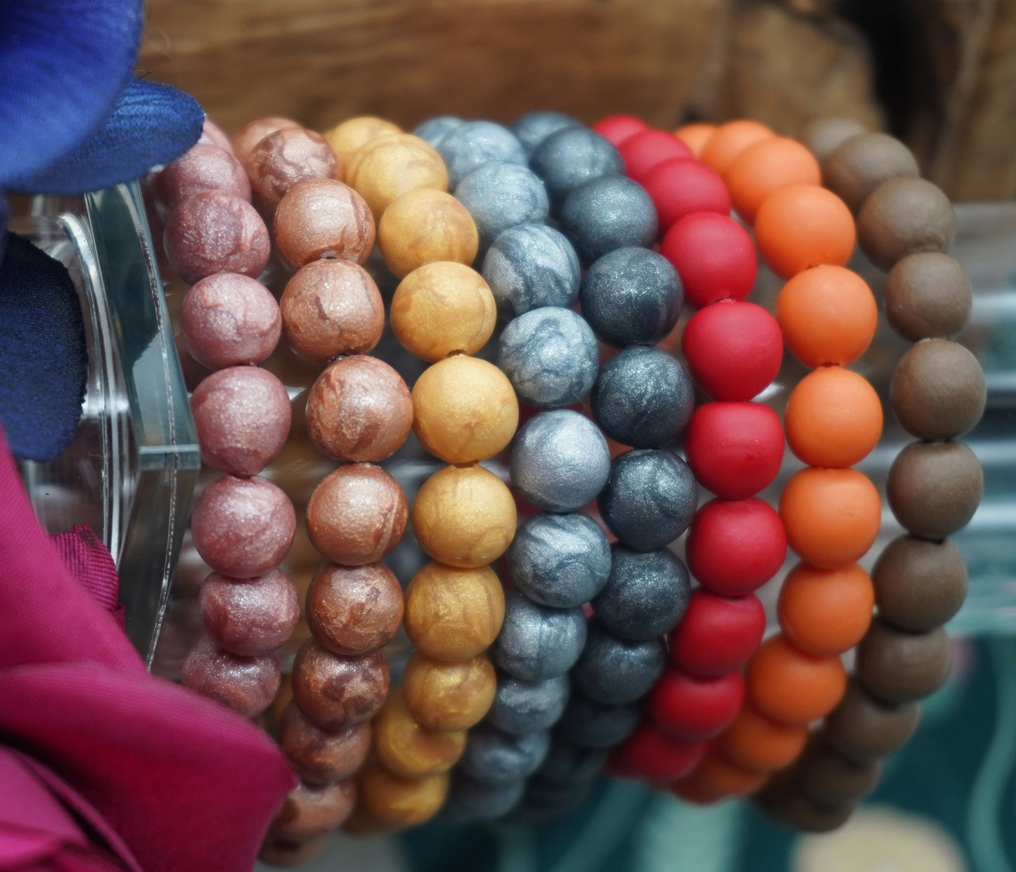 Eight bracelets made of round beads are on a clear display.  Each bracelet is a different color; rose gold, copper, gold, silver, dark grey, red, orange, and dark brown.